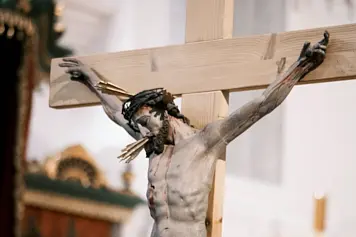 Detailed photograph of a crucified Christ figure on a wooden cross, draped in a white cloth, with a golden crown of thorns.
                                             The cross is adorned with vibrant flowers and greenery, set against a backdrop of a dimly lit church interior.